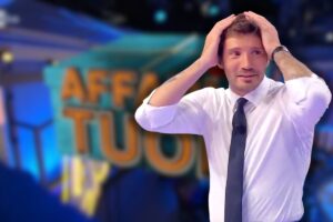logo affari tuoi e stefano de martino con le mani nei capelli